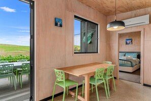 Ein Zimmer mit Holztisch und -stühlen, einem Bett mit zebra-gemustertem Überwurf und einem Fenster mit einer grünen Skulptur, sowie einem Klimagerät.