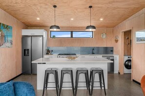 A modern kitchen with a central island, bar stools, a refrigerator, a washing machine, and wooden walls. - Hidden Treasure in Mangawhai Village + EV charger (Mangawhai)