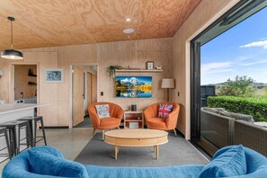 A modern interior with a wooden ceiling, a dining area, two armchairs, a round coffee table, and a large window overlooking greenery. - Hidden Treasure in Mangawhai Village + EV charger (Mangawhai)
