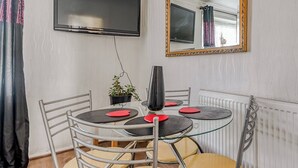 Ein Essbereich mit einem runden Tisch, Stühlen, einer Topfpflanze und einem an der Wand befestigten Fernseher.
