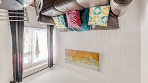 Ein Zimmer mit einem braunen Ledersofa, bunten Kissen, einem Fenster mit Vorhängen und einer Wand mit einem Gemälde.