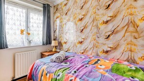 Ein Schlafzimmer mit strukturierter Tapete, einem Bett mit bunter Decke, einem Fenster mit Vorhängen und einem Heizkörper.