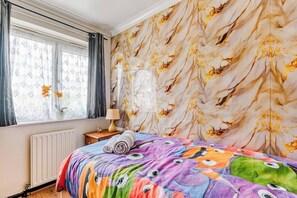 Ein Schlafzimmer mit strukturierter Tapete, einem Bett mit bunter Decke, einem Fenster mit Vorhängen und einem Heizkörper.