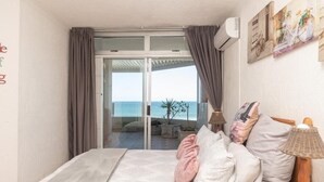 A bedroom with a bed, pillows, a nightstand, a lamp, and a view of the sea through a sliding glass door.