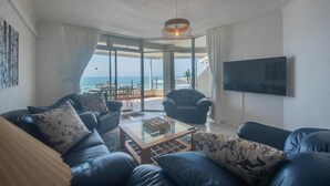 A living room with a sofa set, a coffee table, and a TV. There is a large window with a view of the ocean.
