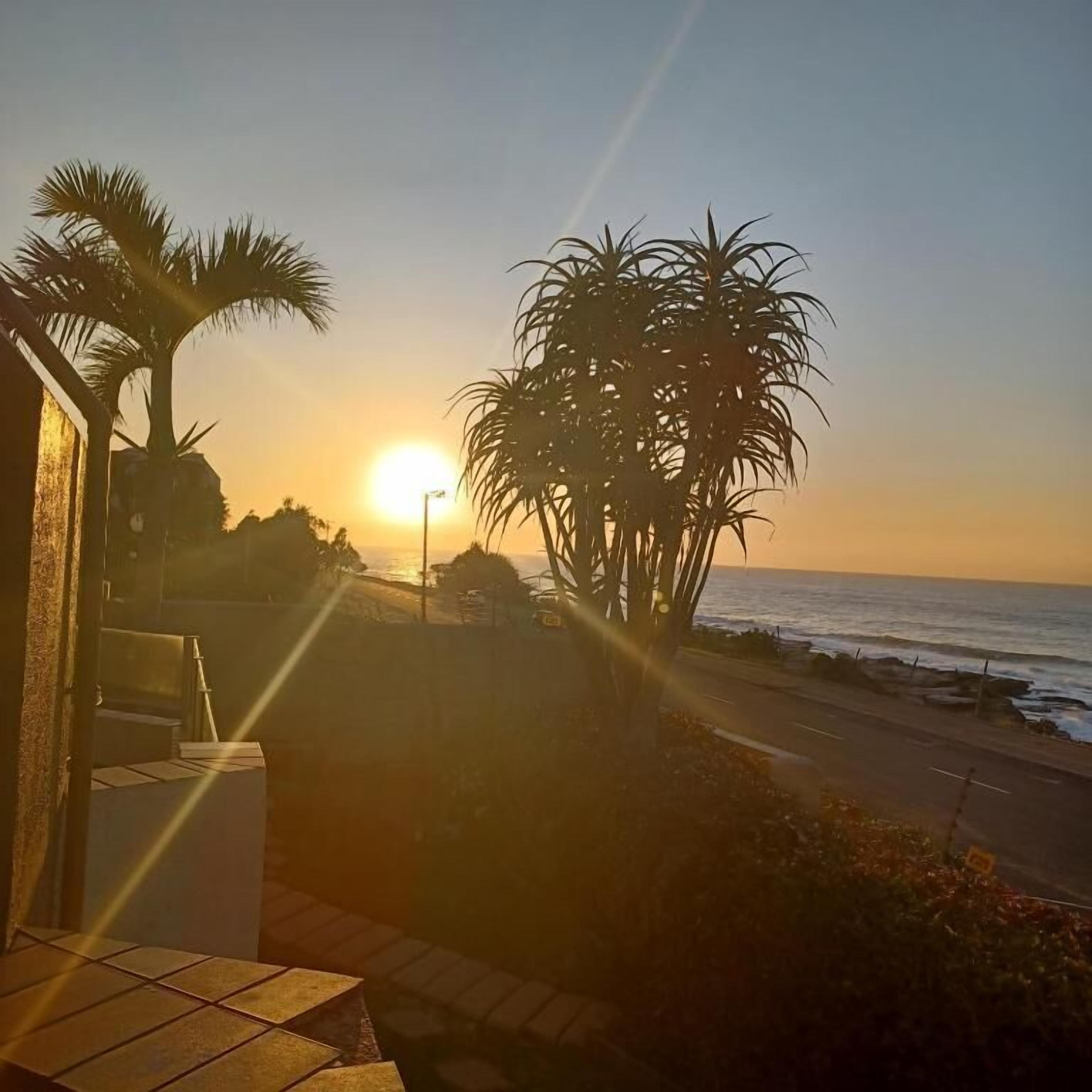Zonsondergang vanaf een balkon met uitzicht op een strand, palmbomen en een weg.