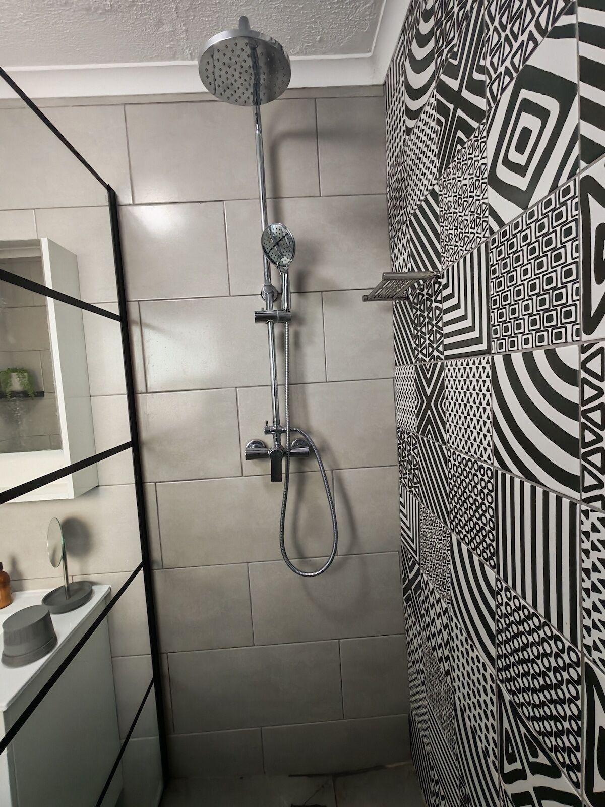 A shower with a handheld showerhead and a handheld hose, a white tiled wall, and a patterned wall.