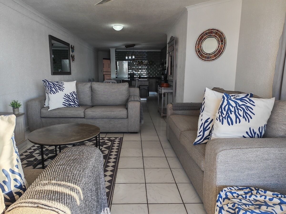 A living room with two sofas, a coffee table, and patterned cushions. A kitchen is visible in the background.