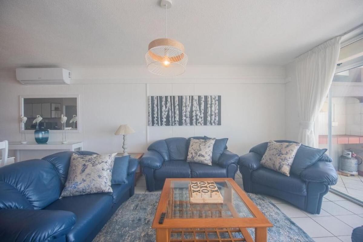 A modern living room with blue leather sofas, a glass coffee table, a hanging light fixture, and a kitchen visible through an open door.