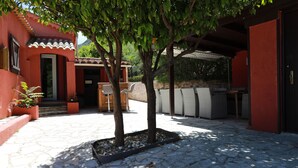 Un pati amb paviment de pedra, un arbre, mobles d'exterior i un edifici amb façana vermella.