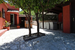 A courtyard with a stone floor, a tree, outdoor furniture, and a building with a red facade. - Villa Oasis Provençale 3 étoiles avec piscine, jacuzzi, terrain de pétanque. (Cavalaire-sur-Mer)