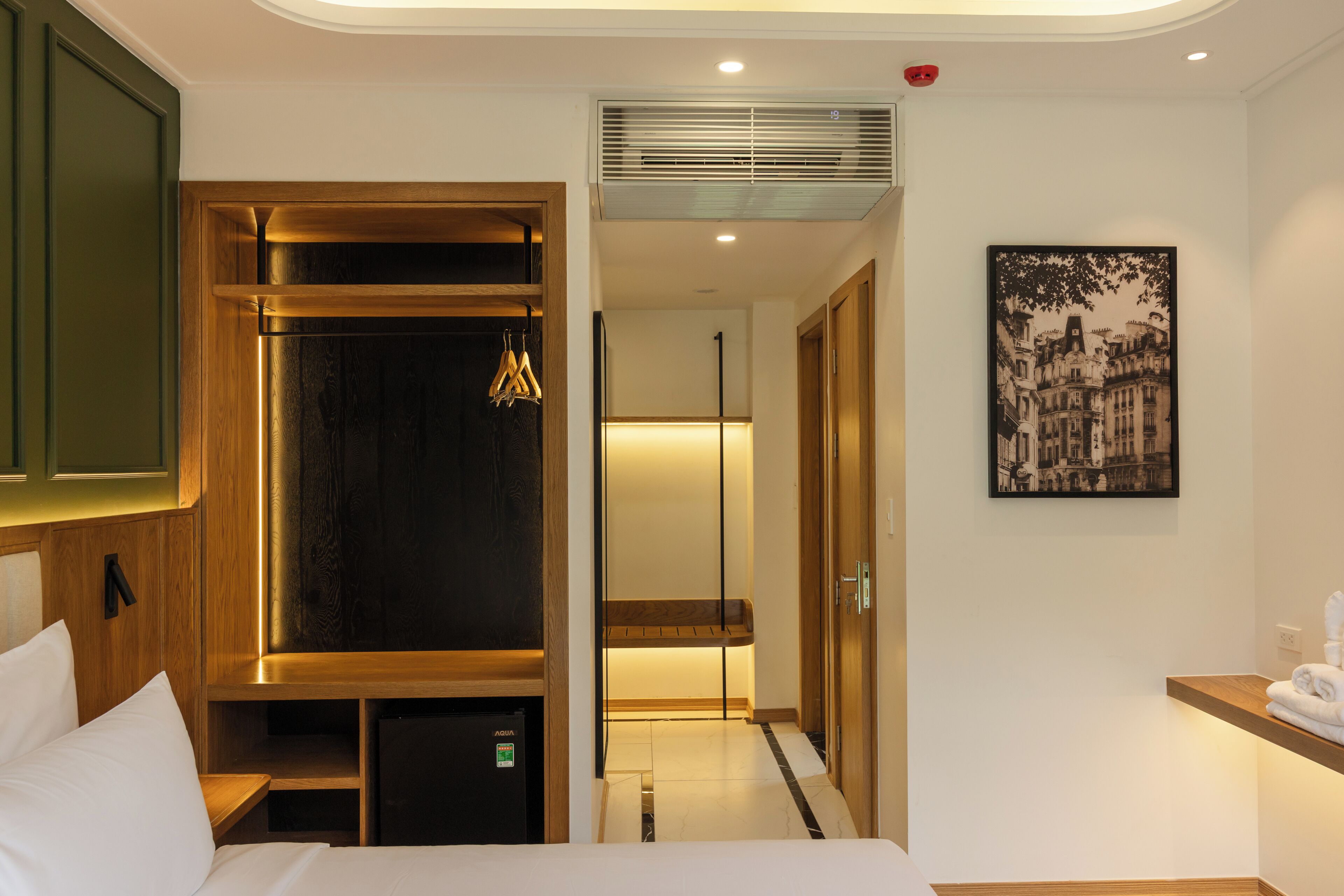 Une chambre d’hôtel comprenant un lit, une armoire, un petit bureau, un miroir, un tableau accroché au mur et un couloir menant à une autre pièce.