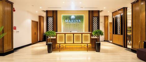 A hotel reception area with a wooden reception desk, two potted plants, and a large illuminated sign.