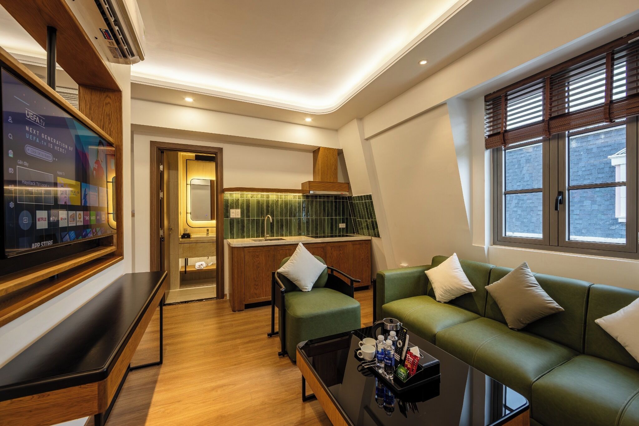Una habitación de hotel moderna con un sofá verde, una mesa de centro, un televisor, una zona de cocina y una ventana con persianas.