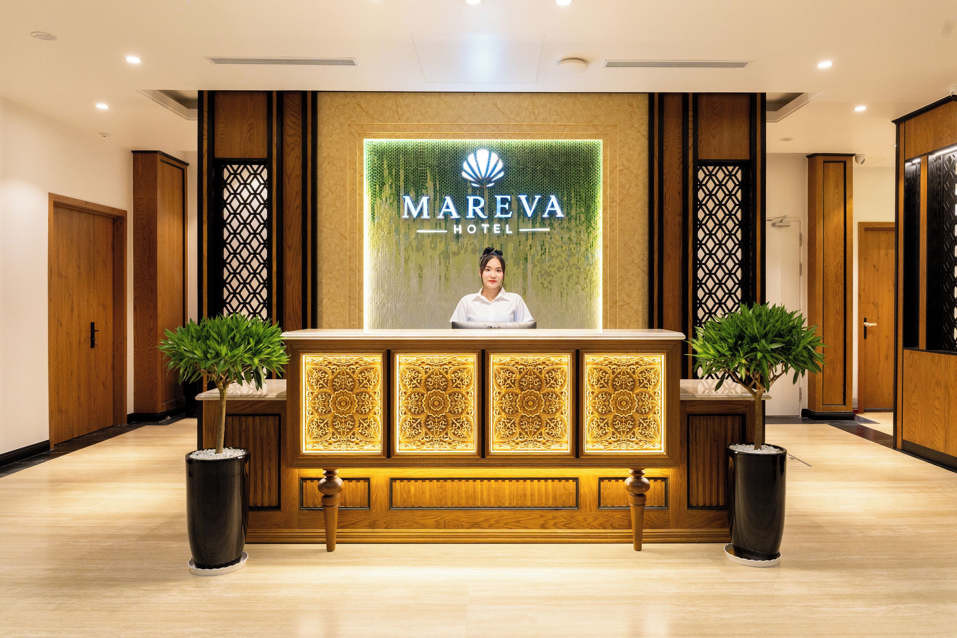 Mostrador de recepción del Hotel Mareva, con una persona detrás, flanqueado por dos plantas en macetas y una pared decorativa con el logotipo del hotel.