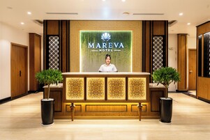 Mostrador de recepción del Hotel Mareva, con una persona detrás, flanqueado por dos plantas en macetas y una pared decorativa con el logotipo del hotel.