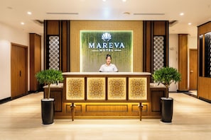 A reception desk at the Mareva Hotel with a person behind it, flanked by two potted plants and a decorative wall with the hotel's logo. - Mareva Khem Beach Hotel (Phu Quoc)