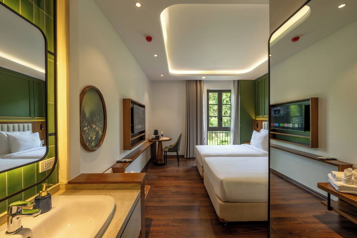 Habitación de hotel con un espejo grande, una cama, un escritorio con una silla, un televisor y una ventana con vistas a zonas verdes.