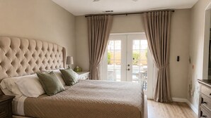Quarto com cabeceira estofada, cama de casal com roupa de cama estampada, mesa de cabeceira com abajur e vista através de uma porta de vidro.