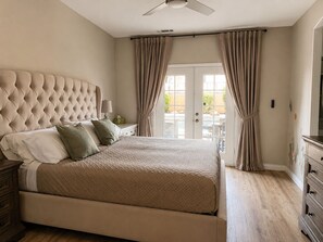 Ein Schlafzimmer mit einem gesteppten Kopfteil, einem Doppelbett mit gemusterten Betttextilien, einem Nachttisch mit Lampe und Blick durch eine Glastür.