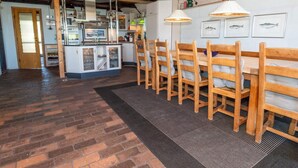 A restaurant interior with wooden chairs, a central dining table, a kitchen area, and a brick floor. - 10 Person Holiday Home in Harboore (Harboore)