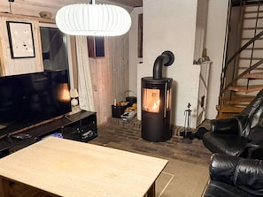 Ein gemütliches Wohnzimmer mit Fernseher, Holztisch, schwarzem Ledersofa, weißer Pendelleuchte und Kaminofen.