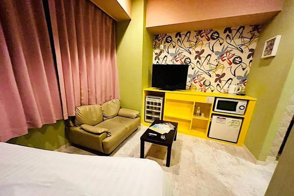 A hotel room with a bed, a sofa, a small table, a TV on a stand, and a minibar.