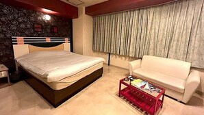 Quarto com cama, mesa de cabeceira, mesa de centro vermelha com revistas, sofá branco e janela com cortinas.