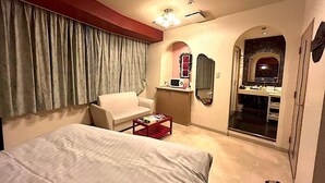 Quarto de hotel com cama, uma cadeira, uma mesinha, um espelho e um banheiro visível através de uma porta em arco.