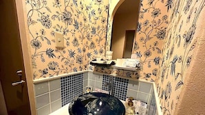 Casa de banho com lavatório preto, papel de parede floral, espelho e uma pequena prateleira com produtos de higiene pessoal.