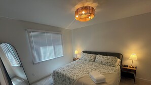 Un dormitorio con una cama, dos lámparas de noche, un despertador y una ventana con persianas, además de una lámpara en el techo.