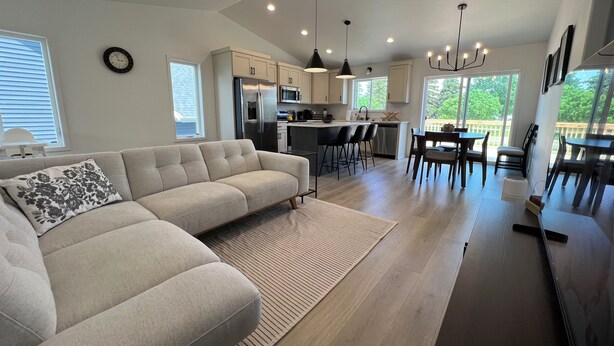 Una sala moderna con sofá, un comedor con mesa y sillas, una cocina equipada y ventanas amplias.