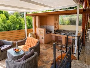 A covered outdoor area with wicker furniture, a grill, and a view of greenery.