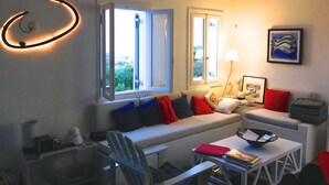 A modern living room with a sofa, armchairs, a coffee table, and a dining table. There are two windows with open curtains, a lamp, and a framed picture on the wall. - A quiet Cycladic-style family home with a pool and an exceptional ocean view. (SIFNOS)
