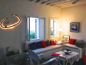 A modern living room with a sofa, armchairs, a coffee table, and a dining table. There are two windows with open curtains, a lamp, and a framed picture on the wall. - A quiet Cycladic-style family home with a pool and an exceptional ocean view. (SIFNOS)