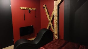 Een kamer met een rode wand, een houten kruisvormige constructie, een zwarte stoel en een bed met een rode spreide.
