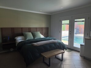 Ein Schlafzimmer mit Bett, Nachttisch, Bank und Blick auf einen Garten mit Pool.