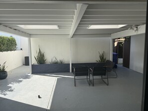 Ein überdachter Patio-Bereich mit Betonboden, einem Tisch mit Stühlen und Topfpflanzen.