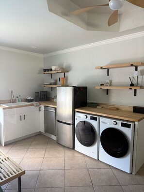 A compact kitchen with a dishwasher, a refrigerator, a washing machine, wooden shelves, and a ceiling fan. - Modern Desert Retreat. Pool & Spa . 3 King Suites. Quiet Upper Cove Location (Cathedral City)