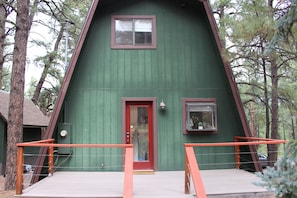 A green A-frame cabin with a red door, two windows, and a wooden deck with orange railings.
