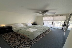 Ein Schlafzimmer mit einem Bett, einem Deckenventilator, einem Nachttisch mit Lampe und einem Fenster mit Vorhängen.