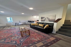 Ein modernes Wohnzimmer mit einer schwarzen Ledersofa, einem gemusterten Teppich, einem Essbereich mit Stühlen, einer Treppe und einem Gemälde an der Wand.