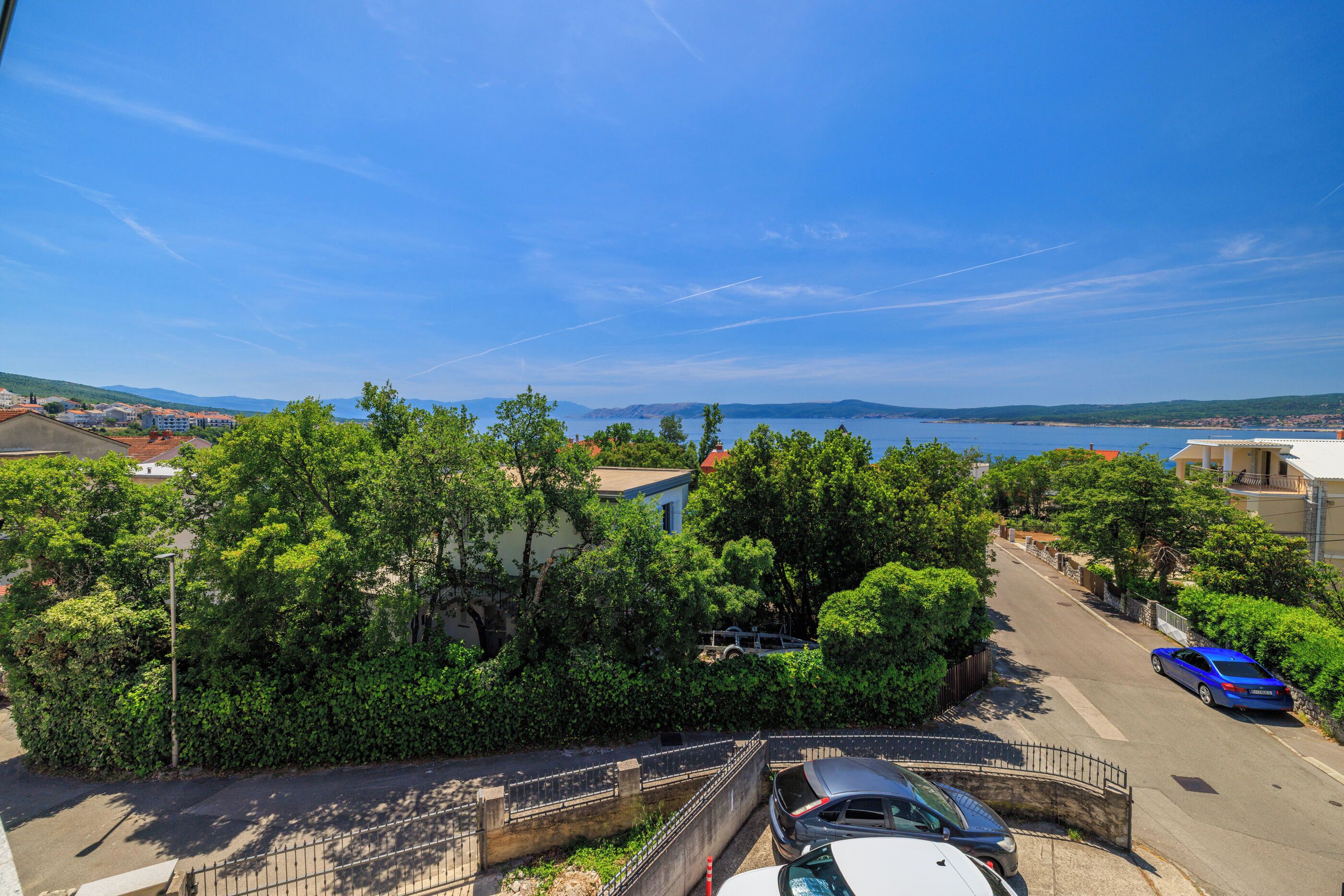 A coastal residential area with a clear view of the sea, a street with parked cars, and lush greenery.