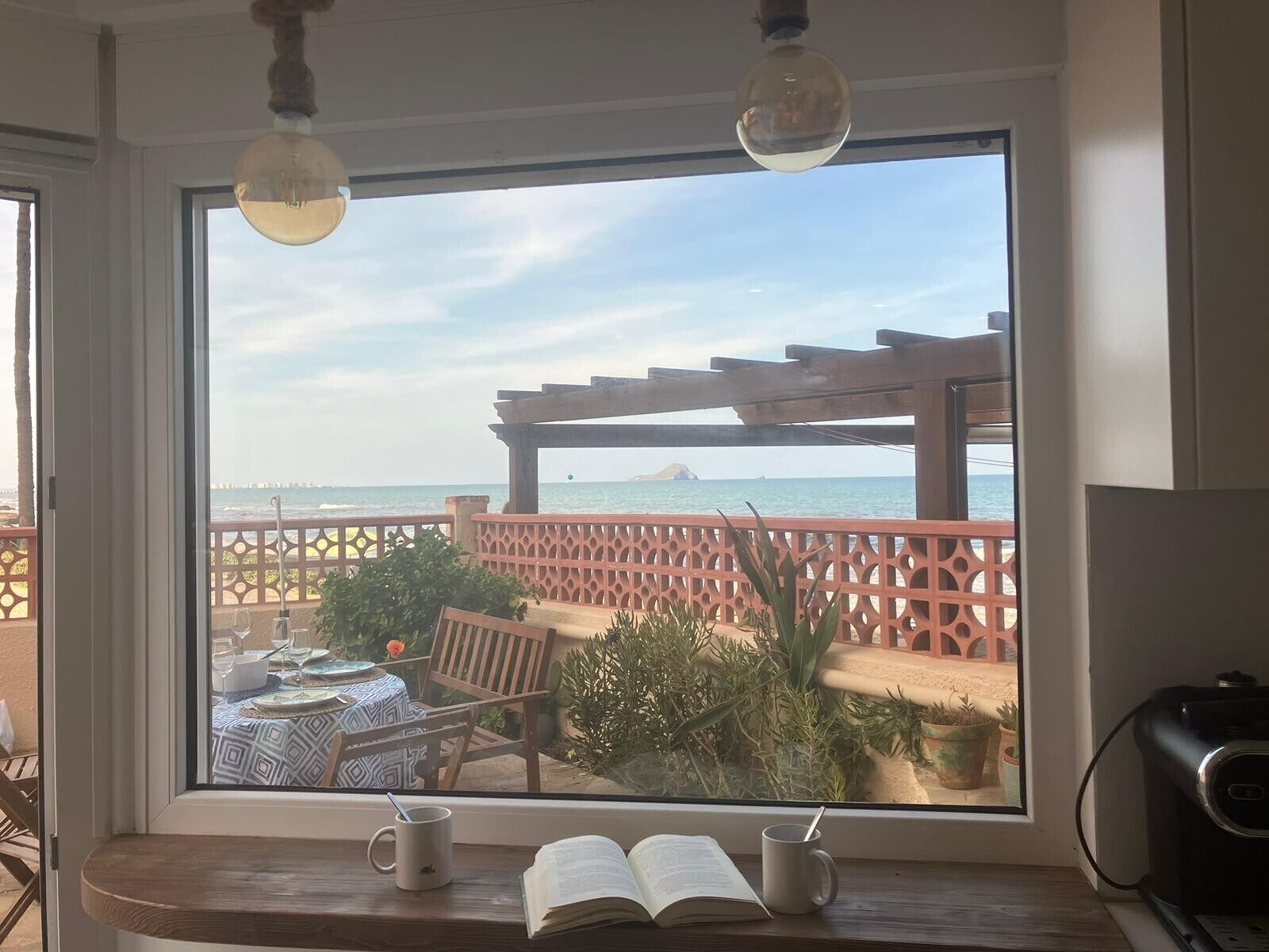 Una habitación con un ventanal grande que da a una playa, una mesa de madera con sillas, y una estantería con libros y una taza.
