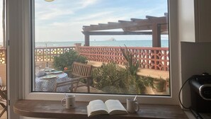 Una habitación con un ventanal grande que da a una playa, una mesa de madera con sillas, y una estantería con libros y una taza.