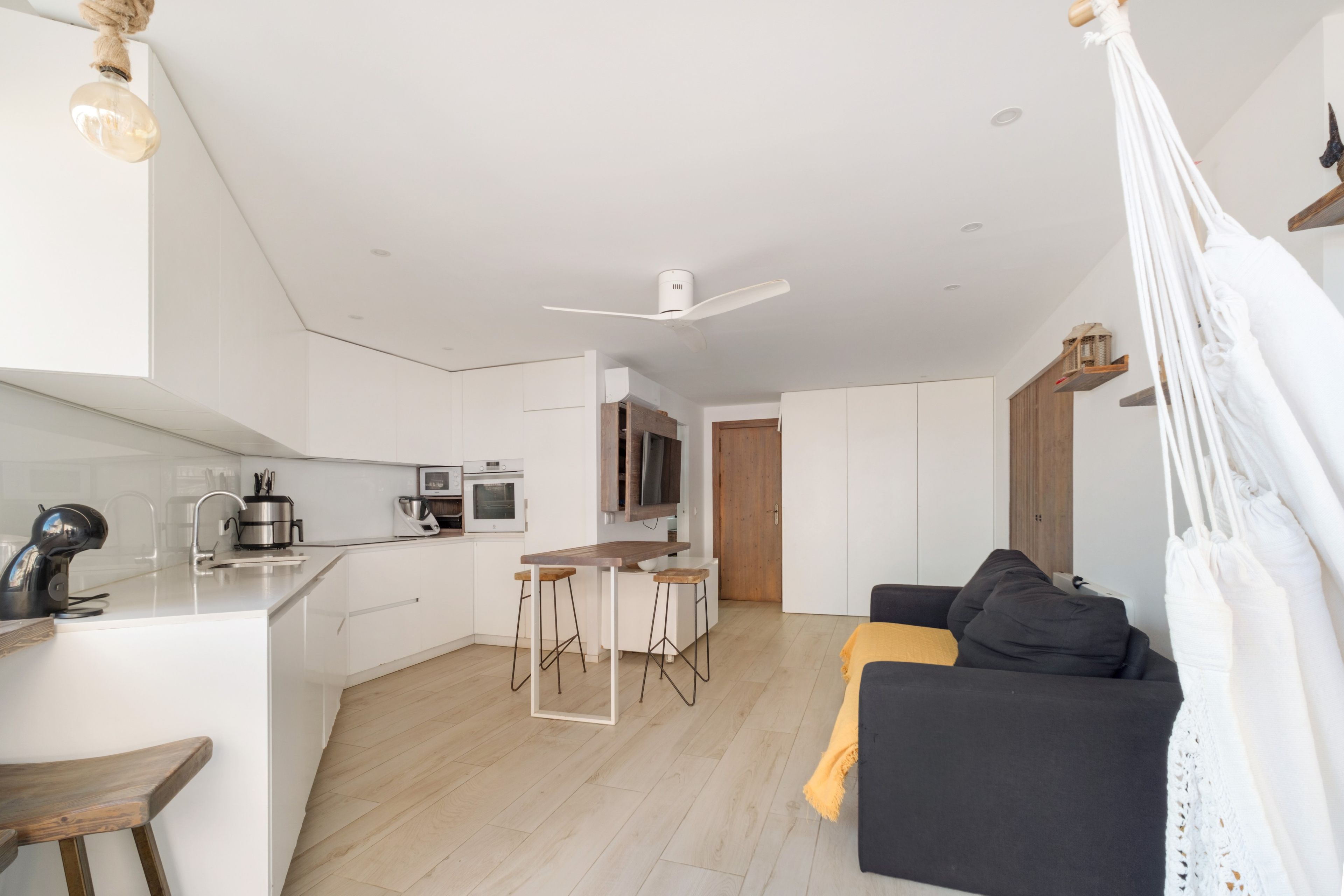 Une cuisine moderne avec des meubles blancs, un îlot central doté de tabourets, un coin repas avec un canapé foncé et un ventilateur de plafond.