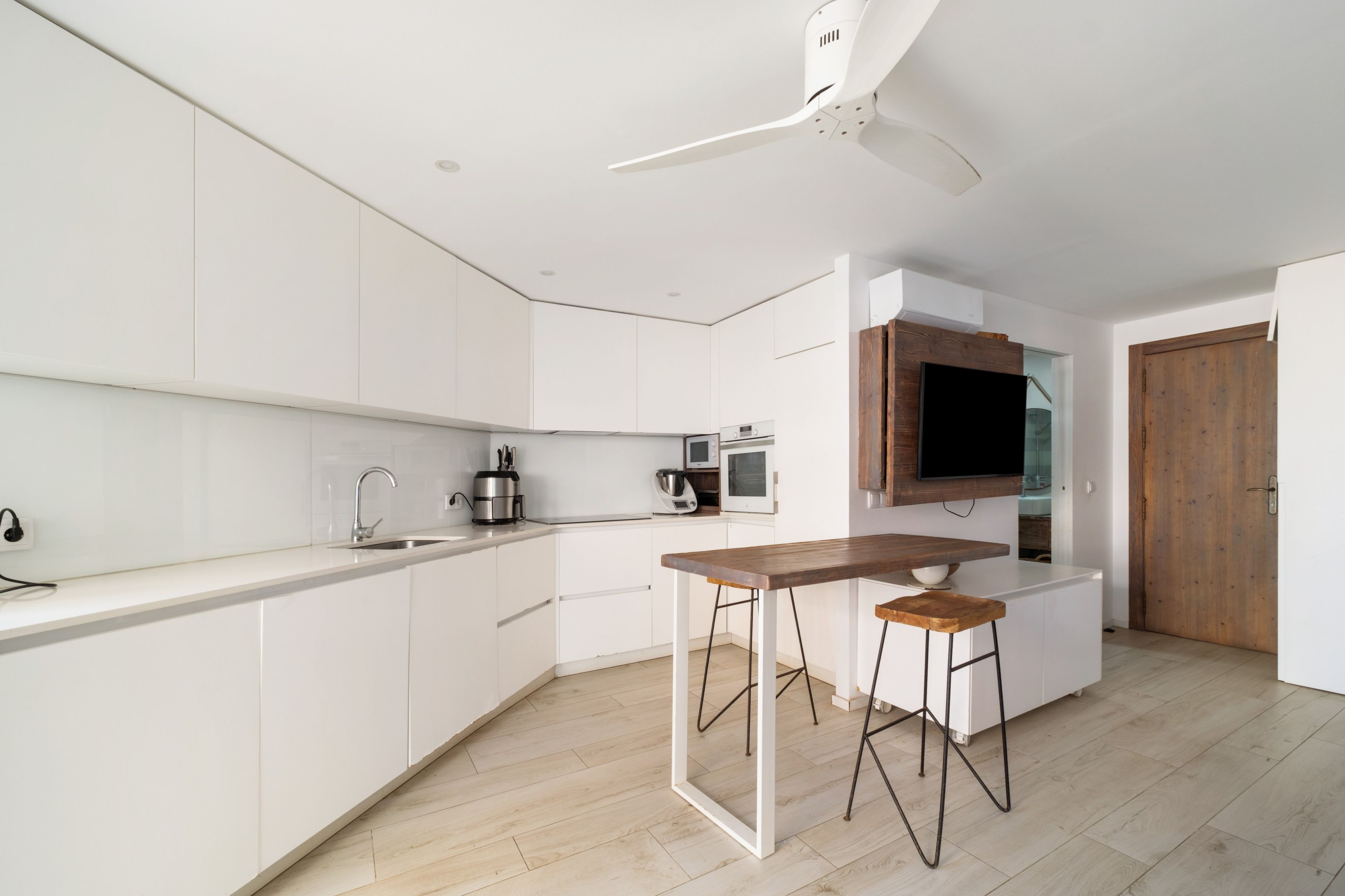 Une cuisine moderne avec des meubles blancs, un îlot central en bois, des tabourets de bar et un ventilateur de plafond.