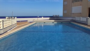 Una piscina con agua cristalina azul, rodeada por una solana empavedada y un edificio con balcones.