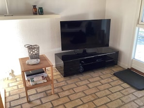 Ein Wohnzimmer mit einem Flachbildfernseher auf einem schwarzen Standfuß, einem kleinen Holztisch mit Korb und Büchern sowie einem Boden mit Ziegeloptik.