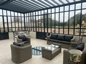 A covered outdoor seating area with wicker furniture and a glass coffee table. - In addition to their room, our guests can enjoy a garden, a terrace, a veranda, and a lounge (Chevanceaux)